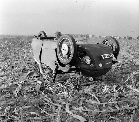 Black and white: three-wheeler lying upside down on stubble, two front wheels in the air, flat-roofed buildings behind.