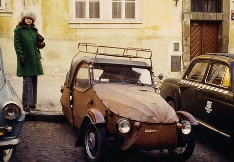 Colour snapshot of a brown fabric-bodied three-wheeler on cobbles between two saloon cars, a woman in a fur hat beside a yellow wall.