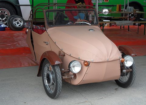 Pink fabric-bodied three-wheeler with brown wings, chrome headlamps and roof hoops, under a gazebo. Opens a larger view