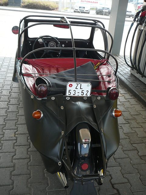 Rear of a black leatherette three-wheeler with tubular hood frame, red seats, Czech number plate and exposed rear wheel at a filling station. Opens a larger view