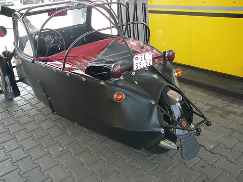 Rear of a black leatherette three-wheeler with roof hoops, red bench seat, single rear wheel and Velorex mudflap. Opens a larger view
