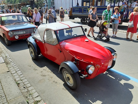 Red three-wheeler with cream fabric hood and black wings parked in a street beside a red Lada, crowds behind. Opens a larger view