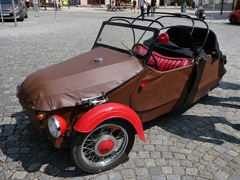 Brown leatherette three-wheeler with bright red front wing, twin horns and black roof hoops, on cobbles. Opens a larger view