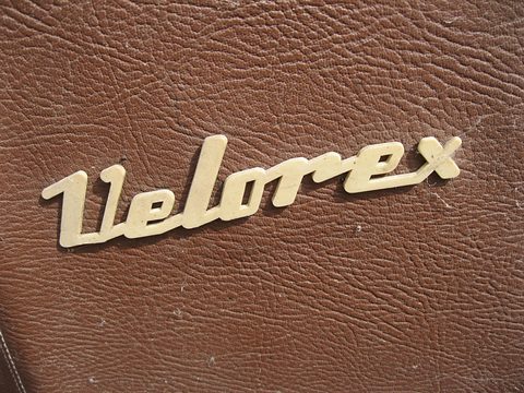 Close-up of a cream plastic Velorex script badge on brown grained artificial leather. Opens a larger view