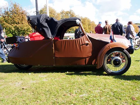 Side of a brown three-wheeler with folded black hood, spoked front wheel and luggage case, on grass among visitors. Opens a larger view