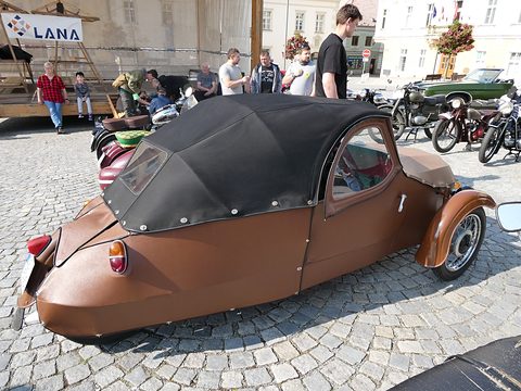 Brown leatherette three-wheeler with black fabric hood, rear lamps and single rear wheel, on a cobbled square. Opens a larger view