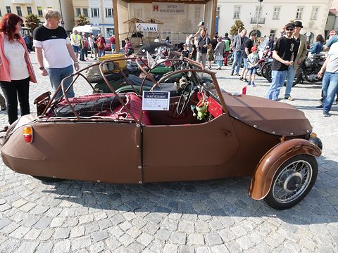 Side of a brown open three-wheeler with roof hoops, red seats and meeting card, on cobbles among visitors. Opens a larger view