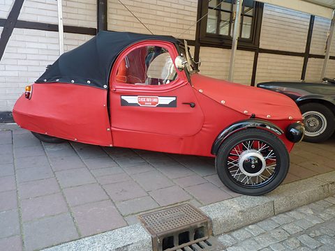 Side of a red three-wheeler with black rear hood, show sticker on the door and spoked front wheel at a kerb. Opens a larger view