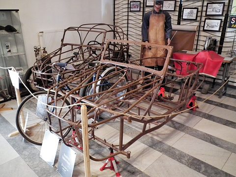 Brown painted tube cage of a three-wheeler on axle stands, with a welder mannequin and sewing machine behind.
