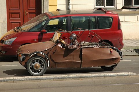 Brown open three-wheeler with roof hoops and spoked wheels passing a parked red people carrier. Opens a larger view