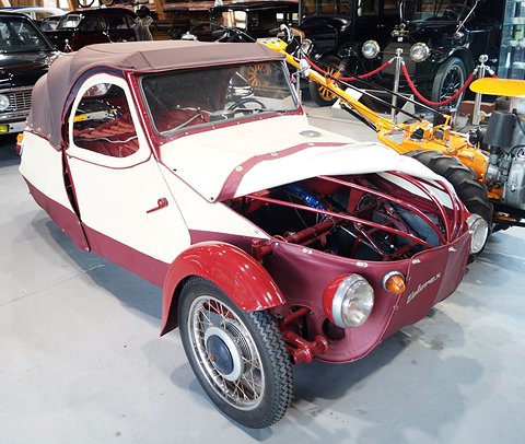 Cream and maroon three-wheeler with front cover folded up showing red tube frame, among vintage cars and a tractor. Opens a larger view