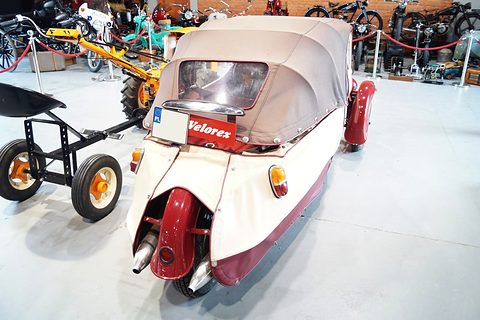 Rear of a cream and maroon three-wheeler with brown hood, single rear wheel and red name panel, in a museum hall. Opens a larger view