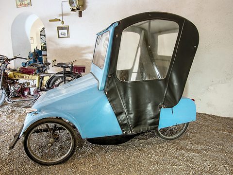Side view of a pale blue voiturette with tall spoked wheels, a black fabric cab with clear windows and veteran motorcycles behind.