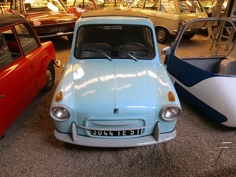 Front of a pale blue small car with two round headlamps and plate 3044 IE 51 between parked cars. Opens a larger view