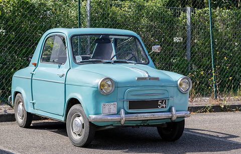 Pale blue small car with a black number plate and rally plaque, parked on gravel before a wire fence and shrubs. Opens a larger view