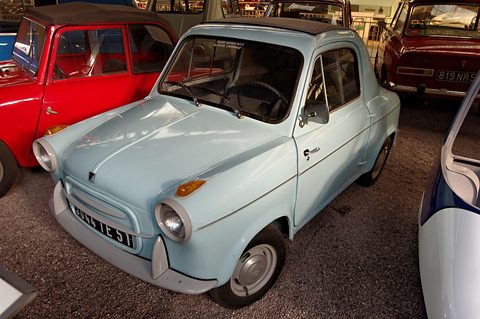 Pale blue small car with orange side indicators on gravel, a red saloon and a white bubble car close by. Opens a larger view