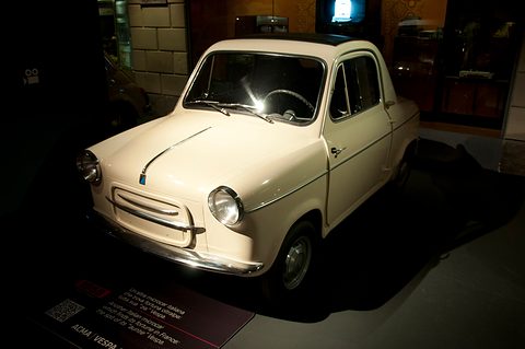 Cream small car spotlit in a dark museum gallery, a placard on the floor in front of it. Opens a larger view
