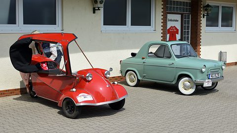 Red three-wheeled roadster with its hood folded open at left, a mint green small car with whitewall tyres parked to its right. Opens a larger view