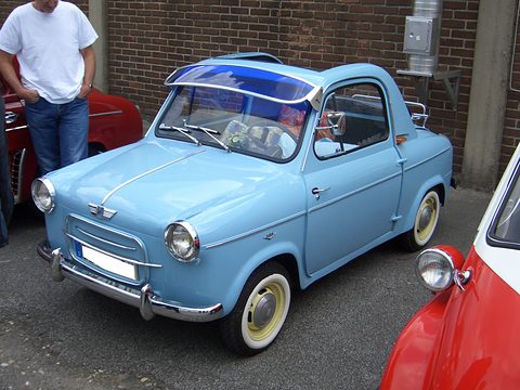 Blue small car with whitewall tyres and a blue sun visor, parked by a brick wall, a red car at its side. Opens a larger view
