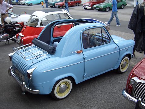 Blue small car with whitewall tyres and a chrome rack on its tail, parked among other cars with people walking by. Opens a larger view