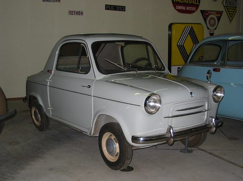 Pale grey small car on a concrete floor in front of old Renault and Vauxhall signs, a blue car beside it. Opens a larger view
