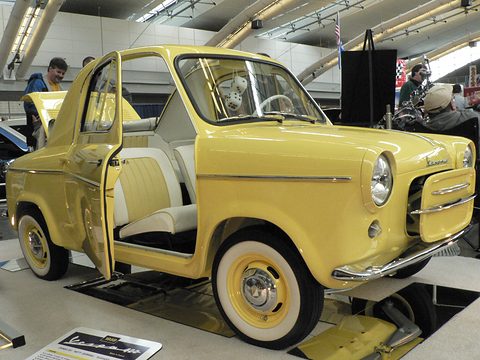Yellow small car with its door swung open on a show stand, pale seats and a white steering wheel inside.