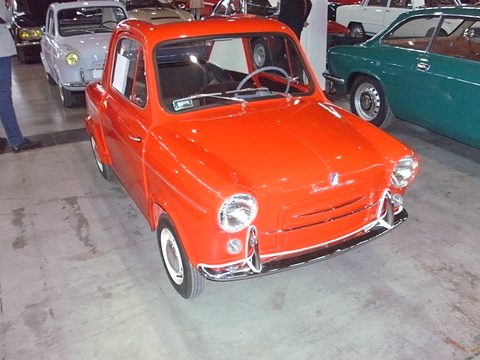 Bright orange small car with chrome bumpers on a stained concrete floor among other classic cars. Opens a larger view