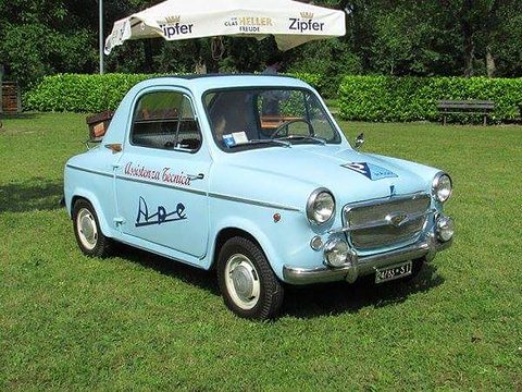 Pale blue small car with a chrome-barred grille, Ape service lettering on its door and a roof rack, on a lawn under a beer umbrella. Opens a larger view
