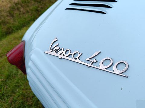 Chrome Vespa 400 script on pale blue paint beside a louvred panel and red tail lamp. Opens a larger view