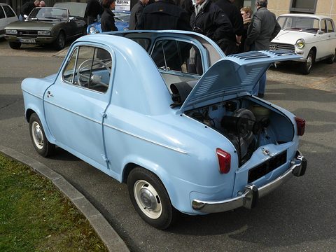 Pale blue small car with its rear lid propped open over the engine, people gathered beside it. Opens a larger view