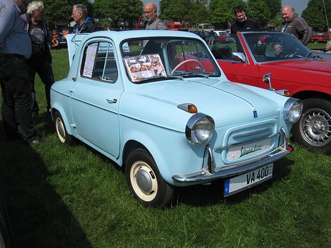 Pale blue small car with front-wing indicators and press cuttings in its window, parked on grass by a red car. Opens a larger view