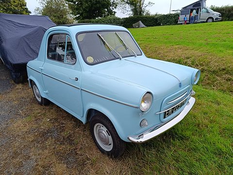 Pale blue small car parked on a grassy verge beside a covered car, a camper van in the distance. Opens a larger view