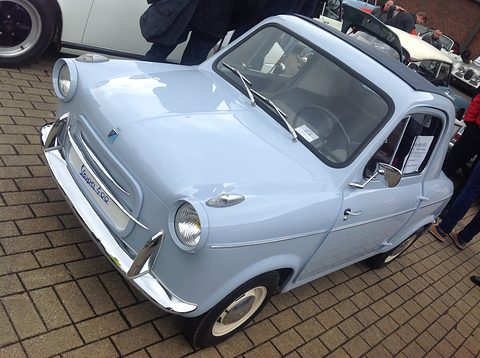 Pale blue small car with Vespa 400 script on its nose and a for-sale card in the window, parked on brick paving. Opens a larger view