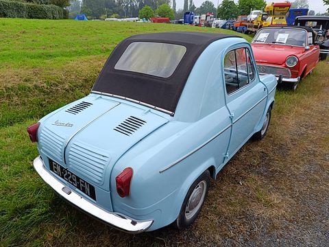 Rear of a pale blue small car with a black roof and louvred engine lid on a verge, a red saloon behind. Opens a larger view