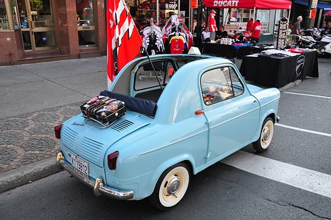 Pale blue small car seen from the rear, roof rolled back and a suitcase strapped to the rack over its engine lid. Opens a larger view