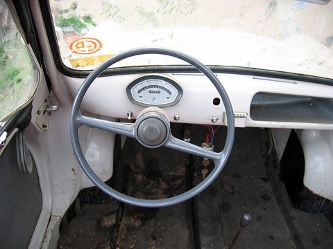 Worn white painted dashboard with a grey steering wheel and round speedometer, a dirty bare floor below. Opens a larger view