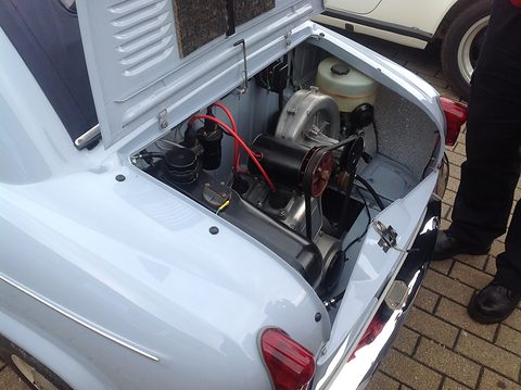 Open rear compartment of a pale blue car with a small engine, cooling fan housing and red ignition leads.
