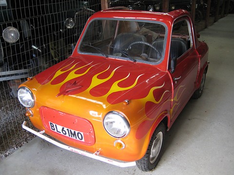 Small red car with yellow and orange flames painted over its nose, parked in a barn beside a wire fence. Opens a larger view