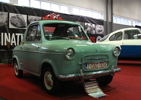 Mint green small car with a Belgian plate on a red carpet, a microcar mural and a two-tone bubble car behind. Opens a larger view