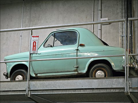 Pale green small car on a steel rack behind railings, a TRAVEE A bay sign in front of its windscreen. Opens a larger view