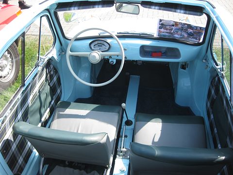 Looking down into a pale blue car interior: two grey seats, a central tunnel with levers and a white steering wheel. Opens a larger view