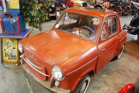 Faded orange small car with a doll in its window, among arcade machines, plants and veteran cars. Opens a larger view