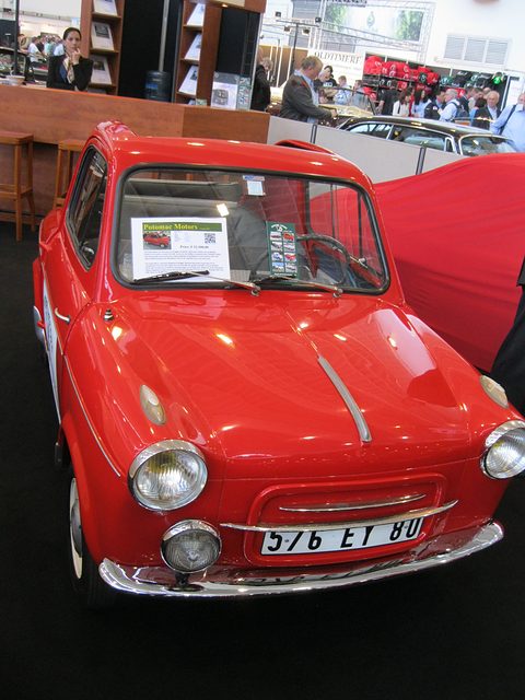 Glossy red small car with plate 576 EY 80 and a sale sheet in its windscreen, on a black show carpet. Opens a larger view