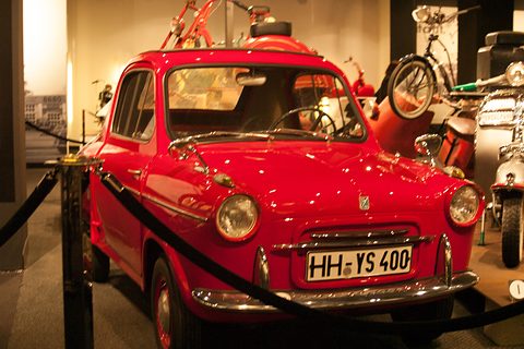 Front of a glossy red small car with round headlamps, a P badge on the nose and a black rope barrier in front. Opens a larger view