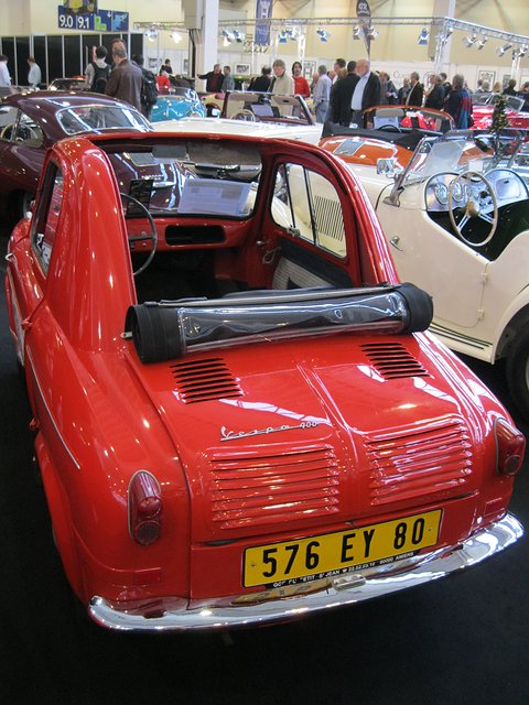 Rear of a red small car with yellow plate 576 EY 80 and its fabric roof rolled forward, crowds behind. Opens a larger view