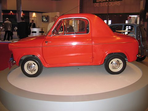 Red small two-door car in profile on a white circular platform in an exhibition hall. Opens a larger view