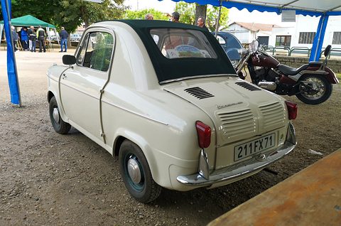 Rear of a cream small car with a dark green fabric roof and red tail lamps, a motorcycle and gazebos behind. Opens a larger view