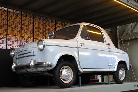White small car seen from below on a raised deck, against a wall of old red and white Belgian number plates. Opens a larger view