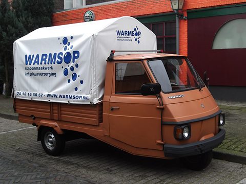 Orange three-wheeled pick-up with a white canvas cover printed with a cleaning firm’s name, parked by a brick building. Opens a larger view