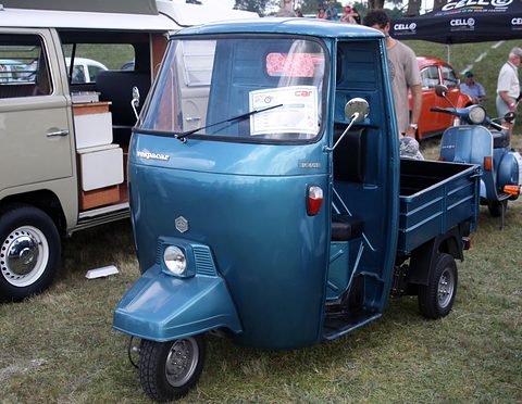 Metallic blue three-wheeled pick-up with a sign on its windscreen, a VW camper and a scooter behind on grass. Opens a larger view